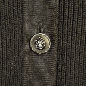 May include: Close-up of a dark gray knit fabric with a decorative button. The button is black with a silver-colored rim and a lion's head design in the center. The fabric has a ribbed texture.