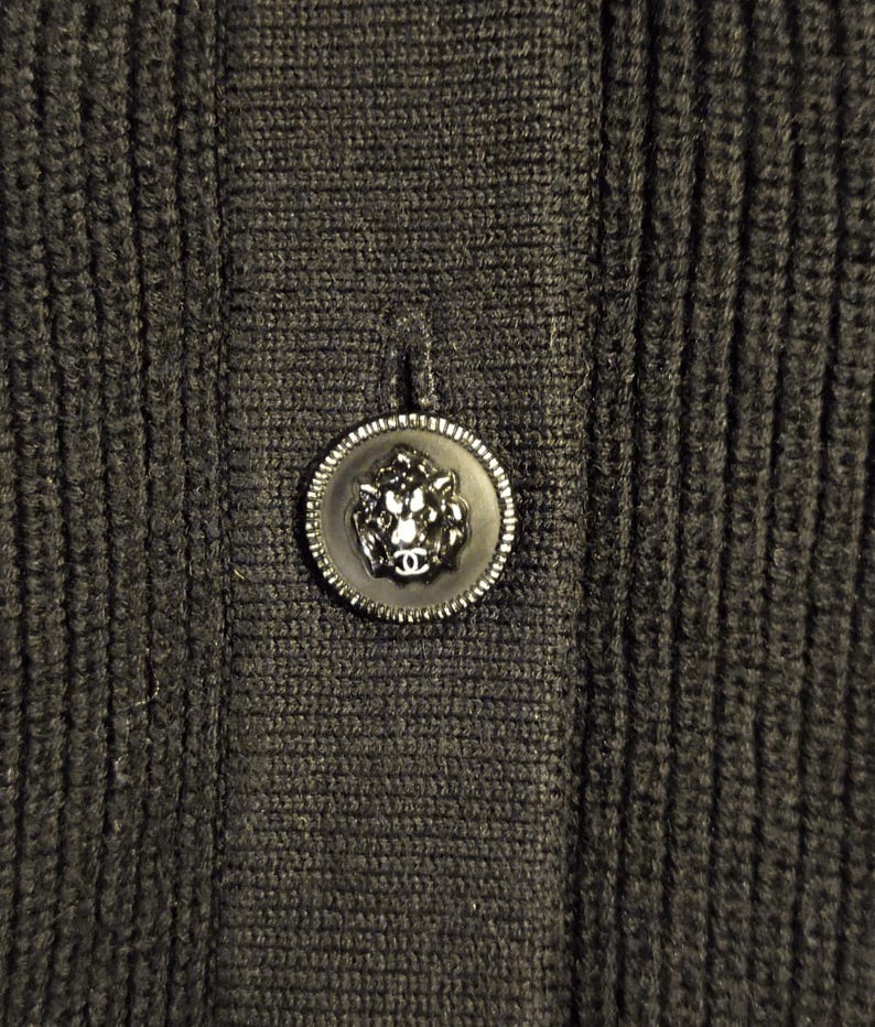 May include: Close-up of a dark gray knit fabric with a decorative button. The button is black with a silver-colored rim and a lion's head design in the center. The fabric has a ribbed texture.