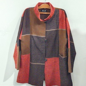 May include: A long-sleeved jacket featuring a patchwork design in shades of rust orange, dark brown, and black. The garment has a high collar, a front button closure with three dark buttons, and a textured fabric construction with varied patterns.