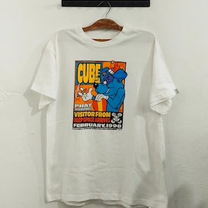 May include: White t-shirt featuring a colorful graphic of a blue dog character with the text "CUBE" and "VISITOR FROM DEEP SPACE ARRIVES FEBRUARY, 1998". The design includes orange, yellow, and red accents.