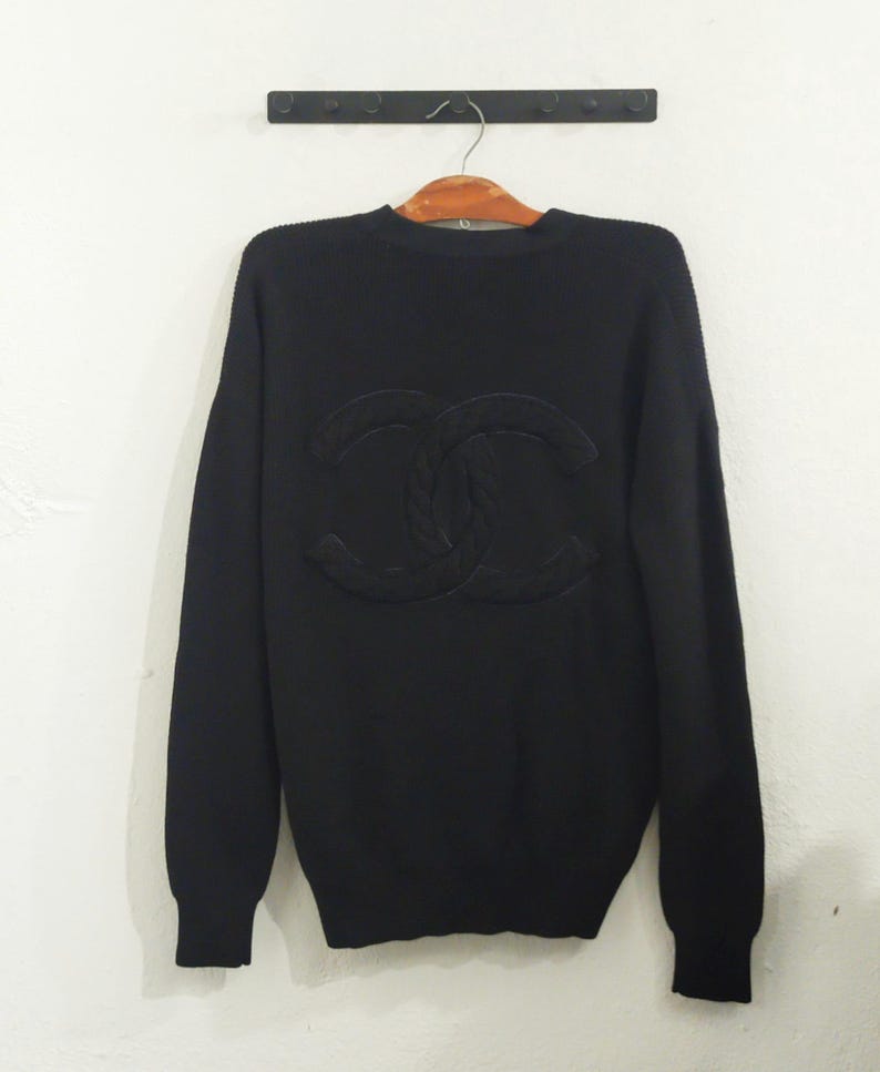May include: Black crew neck sweater with a large, textured logo on the front. The sweater has long sleeves and ribbed cuffs and hem. The logo is a tonal, raised design.