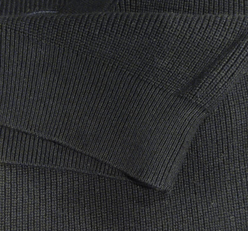 May include: Close-up of a section of a dark gray knit garment, possibly a sweater or jacket. The fabric has a ribbed texture, with visible vertical lines. The sleeve cuff is visible, showing the folded edge and the knit pattern.