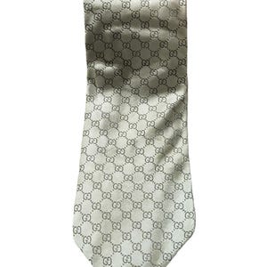 May include: A necktie in a light cream or off-white color featuring a repeating dark gray interlocking GG logo pattern across the entire surface. The fabric has a smooth, satin-like sheen and a pointed tip at the bottom end of the necktie.