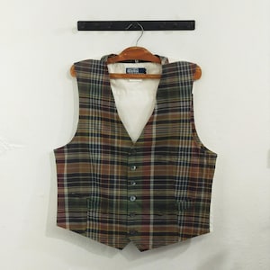 May include: A tartan waistcoat in brown, green, and burgundy, with a V-neck and button-down front. The vest has two front pockets and is hanging on a wooden hanger. The Polo logo is visible on the label.