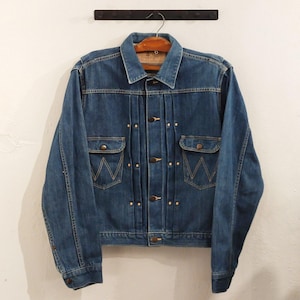 Könnte beinhalten: Blaue Jeansjacke mit Kragen und Knopfverschluss. Die Jacke hat zwei Brusttaschen mit einem markanten 'W'-Design und messingfarbenen Knöpfen. Die Jacke hängt an einem Holzbügel vor einer weißen Wand.