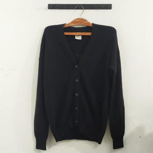 May include: A black cardigan sweater with a V-neck and button-down front. The sweater is hanging on a wooden hanger against a white wall. The sweater has long sleeves and a classic design.