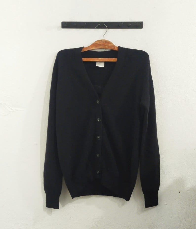 May include: A black cardigan sweater with a V-neck and button-down front. The sweater is hanging on a wooden hanger against a white wall. The sweater has long sleeves and a classic design.