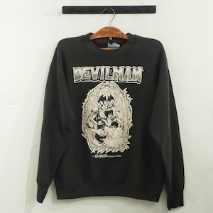 May include: Black sweatshirt featuring the text "DEVILMAN" in a white and green font above a detailed graphic. The graphic depicts several figures in a dynamic pose. The sweatshirt is hanging on a wooden hanger.