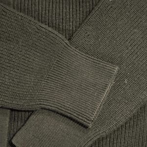 May include: Close-up of a dark green cardigan sweater. The ribbed knit fabric is visible on the sleeve and body. The sleeve cuff is a solid knit. A button is visible on the upper left side of the sweater.