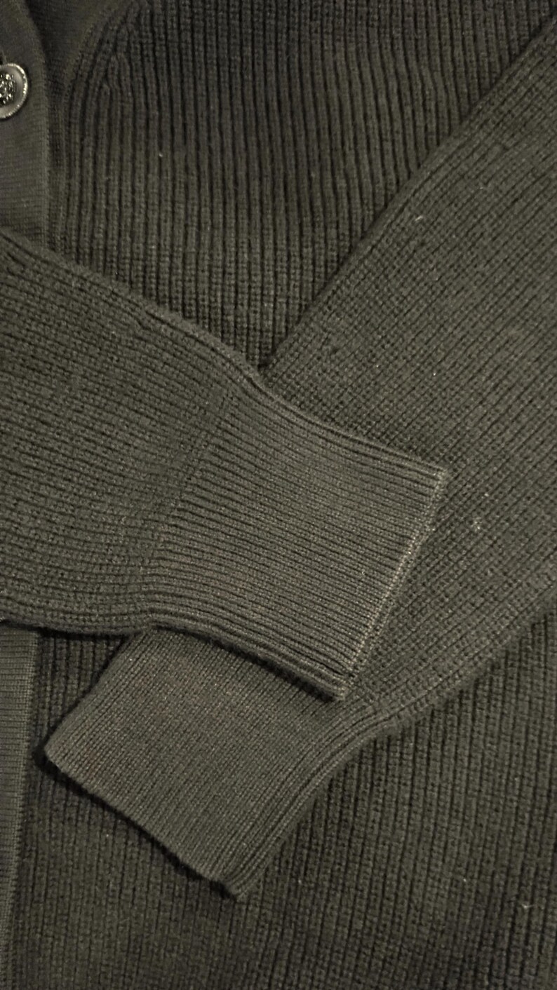 May include: Close-up of a dark green cardigan sweater. The ribbed knit fabric is visible on the sleeve and body. The sleeve cuff is a solid knit. A button is visible on the upper left side of the sweater.