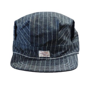 Pode incluir: Boné de ganga azul com riscas brancas verticais finas. O boné tem uma aba plana e uma borda branca na borda. Uma etiqueta retangular na frente diz "Red Cloud SHERALL MFG CO. TRADE MARK".