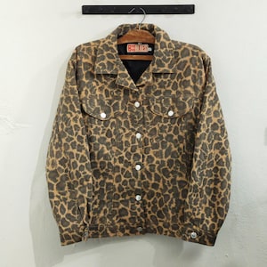 May include: A brown and tan leopard print denim jacket with silver buttons. The jacket has two chest pockets and a black collar. The label reads "BY BLUES". The jacket is hanging on a wooden hanger.