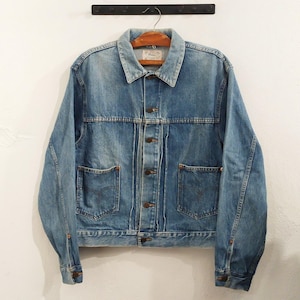 May include: A vintage blue denim jacket with a classic design. The jacket has a button-up front, two chest pockets, and two side pockets. The jacket is hanging on a wooden hanger against a white wall.
