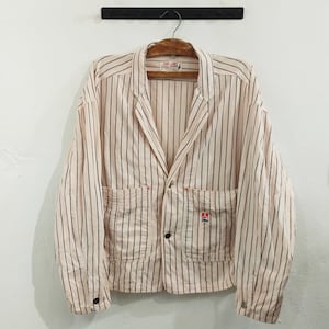 May include: A light pink blazer with vertical black stripes, two front pockets, and black buttons. The blazer is hanging on a wooden hanger. A small logo is visible on the right pocket.