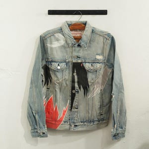 Puede incluir: Chaqueta vaquera azul claro con obras de arte pintadas a mano. La chaqueta presenta un diseño desgastado con rasgaduras y desgarros, y está adornada con elementos pintados en negro, rojo y gris. La chaqueta está colgada en una percha de madera.