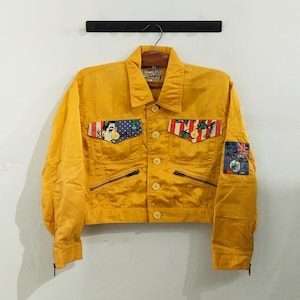 Puede incluir: Chaqueta amarilla mostaza con parches decorativos. La chaqueta tiene cuello, cierre de botones y dos bolsillos con cremallera. Los parches presentan una imagen de estilo cómic, un diseño rojo, blanco y azul y una bandera de la Union Jack.