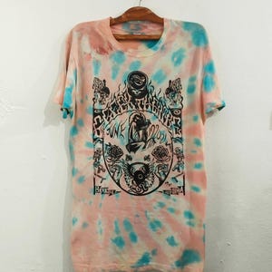 May include: A tie-dye t-shirt in shades of pink and blue, hanging on a wooden hanger. The shirt features a black graphic design with text that reads "Sunk 19202" and "Berkeley California".