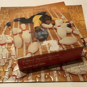 May include: A red and white box with a monkey bowling. The box says "Throw yourself into everything you do!" and "A Mini Jigsaw Puzzle 7 in. x 7 in. / Over 70 Pieces" with the Springbok logo. The box is sitting on top of a completed jigsaw puzzle of a bowling alley.