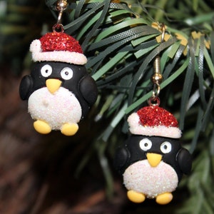 K&ouml;nnte beinhalten: Ein Paar H&auml;ngeohrringe mit einem Pinguin, der einen roten und wei&szlig;en Weihnachtsmann-Hut tr&auml;gt. Der Pinguin ist aus wei&szlig;em und schwarzem Polymerton mit Glitzerakzenten gefertigt.