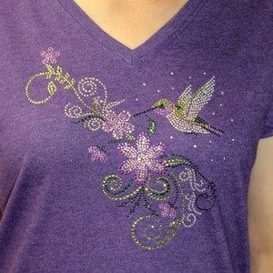 Puede incluir: Camiseta morada con un colibrí y un diseño floral de pedrería.