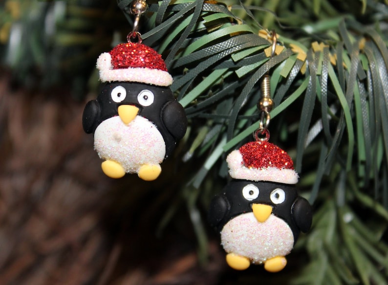 K&ouml;nnte beinhalten: Ein Paar H&auml;ngeohrringe mit einem Pinguin, der einen roten und wei&szlig;en Weihnachtsmann-Hut tr&auml;gt. Der Pinguin ist aus wei&szlig;em und schwarzem Polymerton mit Glitzerakzenten gefertigt.