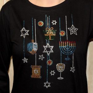 Puede incluir: Camiseta negra de manga larga con un diseño festivo de Janucá. La camiseta presenta adornos de pedrería brillantes de una menorá, una Estrella de David, un dreidel y otros símbolos festivos. El diseño incluye detalles en azul, dorado, rojo y plateado.