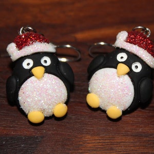 K&ouml;nnte beinhalten: Ein Paar H&auml;ngeohrringe mit einem Pinguin, der einen roten und wei&szlig;en Weihnachtsmann-Hut tr&auml;gt. Der Pinguin ist aus wei&szlig;em und schwarzem Polymerton mit Glitzerakzenten gefertigt.