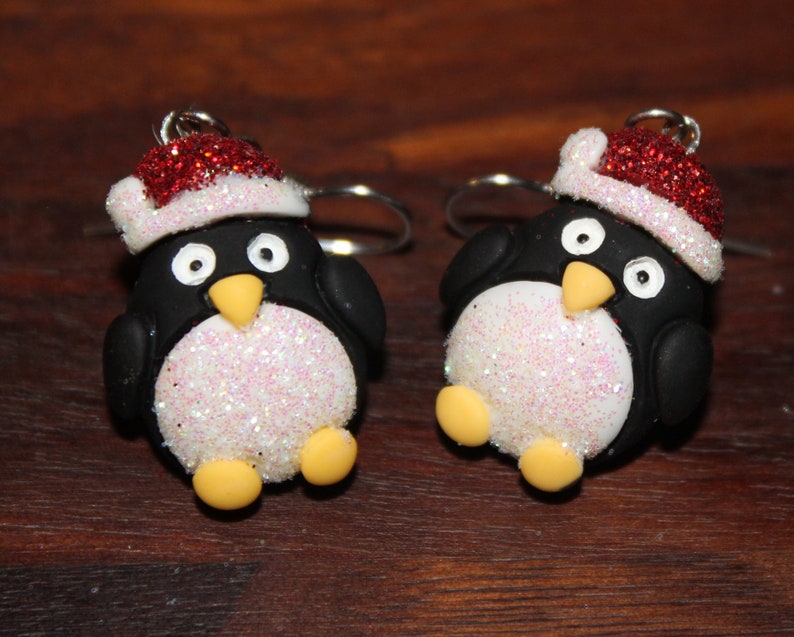 K&ouml;nnte beinhalten: Ein Paar H&auml;ngeohrringe mit einem Pinguin, der einen roten und wei&szlig;en Weihnachtsmann-Hut tr&auml;gt. Der Pinguin ist aus wei&szlig;em und schwarzem Polymerton mit Glitzerakzenten gefertigt.