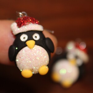 K&ouml;nnte beinhalten: Ein Paar H&auml;ngeohrringe mit einem Pinguin, der einen roten und wei&szlig;en Weihnachtsmann-Hut tr&auml;gt. Der Pinguin ist aus wei&szlig;em Glitzer und schwarzem und wei&szlig;em Ton gefertigt.