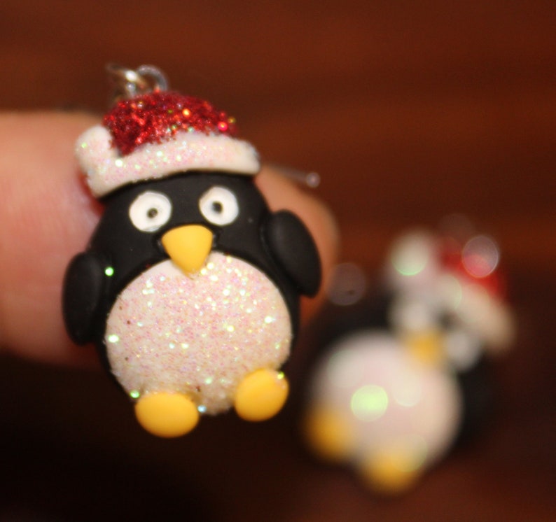 K&ouml;nnte beinhalten: Ein Paar H&auml;ngeohrringe mit einem Pinguin, der einen roten und wei&szlig;en Weihnachtsmann-Hut tr&auml;gt. Der Pinguin ist aus wei&szlig;em Glitzer und schwarzem und wei&szlig;em Ton gefertigt.