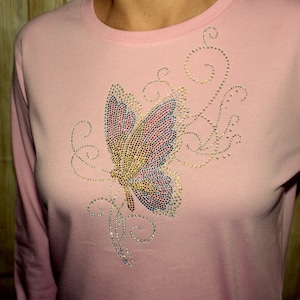 May include: A pink long-sleeved shirt with a sparkly butterfly design. The butterfly is made of rhinestones and has a light blue, pink, and yellow color scheme.