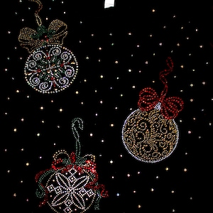 Puede incluir: Tela negra adornada con tres dise&ntilde;os de adornos navide&ntilde;os. Cada adorno est&aacute; adornado con brillantes pedrer&iacute;a de varios colores, incluyendo rojo, verde y dorado. Los adornos est&aacute;n acentuados con lazos y una dispersi&oacute;n de peque&ntilde;os puntos brillantes.