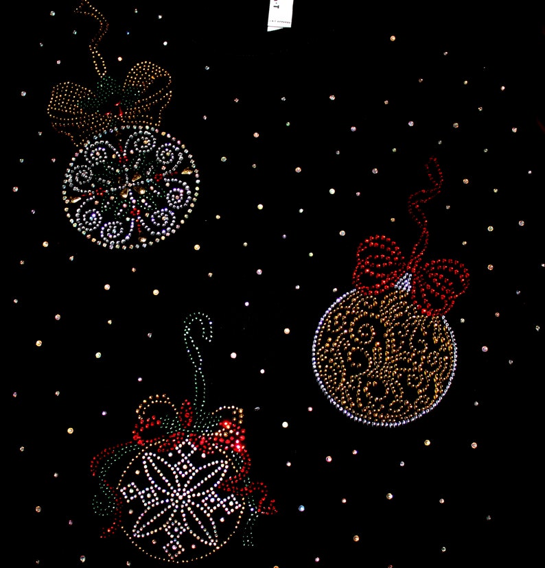 Puede incluir: Tela negra adornada con tres dise&ntilde;os de adornos navide&ntilde;os. Cada adorno est&aacute; adornado con brillantes pedrer&iacute;a de varios colores, incluyendo rojo, verde y dorado. Los adornos est&aacute;n acentuados con lazos y una dispersi&oacute;n de peque&ntilde;os puntos brillantes.