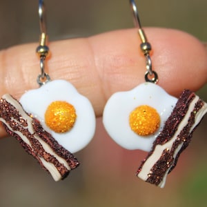 May include: A pair of dangle earrings featuring a white fried egg with a yellow yolk and a brown and white piece of bacon. The earrings are made of polymer clay and have a silver hook.