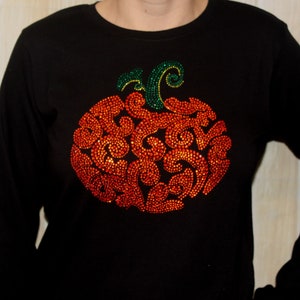 May include: Black long-sleeved shirt with a large, sparkly orange pumpkin design. The pumpkin is made of rhinestones and has a green stem.