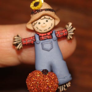 May include: A small scarecrow figurine featuring a tan hat with a yellow sunflower, a red and black plaid shirt, and blue overalls. The figure has a smiling face and is positioned behind a round, glittery orange pumpkin with a black stem.
