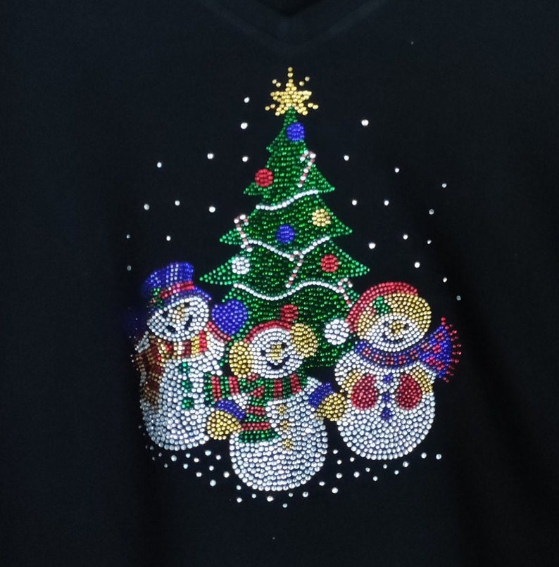 May include: A black Christmas jumper featuring a green, red and blue Christmas tree and three snowmen made of rhinestones.