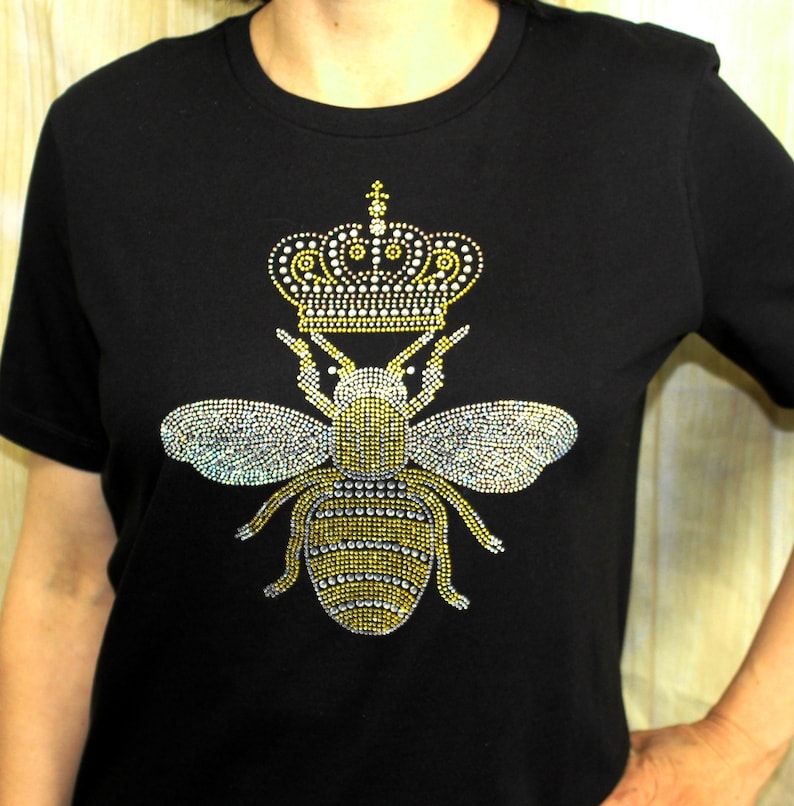 Peut inclure: T-shirt noir avec un motif d'abeille en strass. L'abeille porte une couronne.