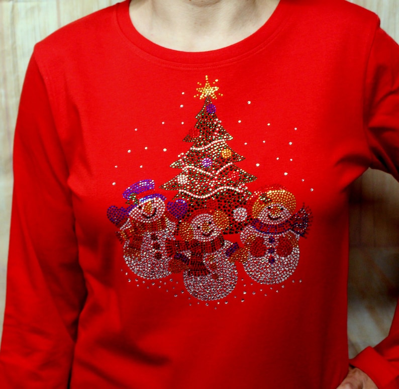 May include: Red long-sleeved shirt featuring a Christmas tree and three snowmen design made of rhinestones.