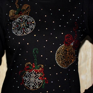 Puede incluir: Camiseta negra de manga larga adornada con tres dise&ntilde;os de bolas de Navidad. Los adornos est&aacute;n adornados con brillantes pedrer&iacute;a en varios colores, incluyendo rojo, dorado y plateado. La pedrer&iacute;a dispersa crea un efecto estrellado.