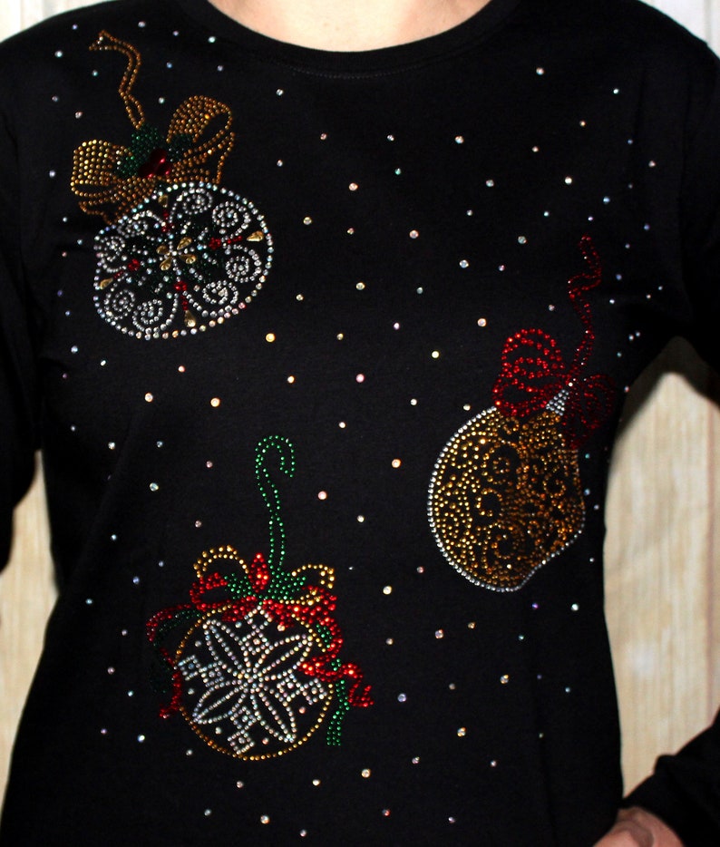 Puede incluir: Camiseta negra de manga larga adornada con tres dise&ntilde;os de bolas de Navidad. Los adornos est&aacute;n adornados con brillantes pedrer&iacute;a en varios colores, incluyendo rojo, dorado y plateado. La pedrer&iacute;a dispersa crea un efecto estrellado.