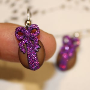 May include: A pair of dangle earrings with a brown base and a purple glitter bow. The bow is made of a sparkly material and has a 3D effect.
