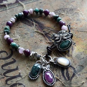 May include: A beaded bracelet featuring a mix of green, pink, and purple gemstones. The bracelet has a silver dragonfly clasp and several dangling charms with gemstones. The bracelet is on a vintage-style background.