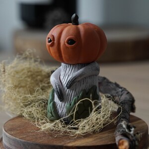 Op de afbeelding: Een klein, handgemaakt beeldje van een wezen met een pompoenhoofd, een wit lichaam en groene bladeren. Het beeldje staat op een houten voet met bruin mos.