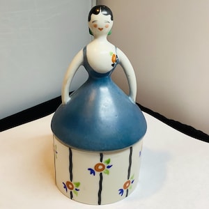 May include: A vintage ceramic figurine of a woman in a blue dress. The figurine has a white torso with a blue dress and a round base. The base is decorated with vertical black stripes and floral designs in orange, blue, and green.