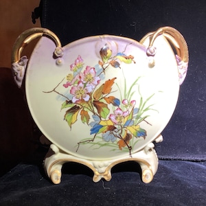 May include: A white porcelain vase with gold accents and floral designs. The vase has two handles and three feet. The floral design features pink, yellow, and blue flowers with green leaves.