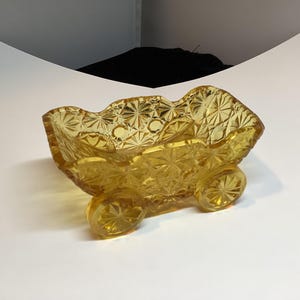 May include: An antique amber glass dish with a starburst design. The dish features a scalloped edge and is supported by three small, rounded feet. The glass has a textured, diamond-like pattern throughout. The dish is approximately 15 cm long.