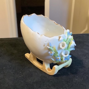 Puede incluir: Un cuenco decorativo de porcelana blanca en forma de huevo sobre una base de trineo, adornado con delicados detalles florales azules y blancos. El borde está adornado con oro. Una pieza encantadora para exhibir o pequeños objetos.