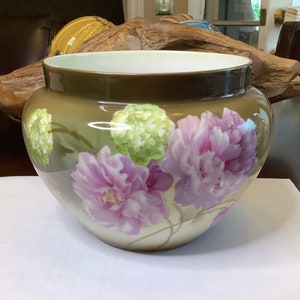 Könnte beinhalten: Ein brauner Keramiktopf mit einem Blumendesign mit rosa und grünen Blumen. Der Topf hat einen breiten, runden Boden und einen leicht ausgestellten Rand.