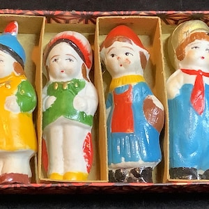 May include: A set of four vintage ceramic figurines in a decorative box. Each figurine is hand-painted with bright colors, including yellow, blue, green, and red. The figures are dressed in various outfits, suggesting a theme of traditional or historical characters.